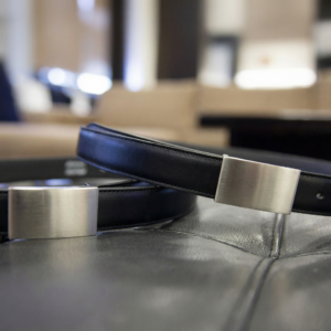 Two black leather belts featuring brushed silver buckles, angled on a dark textured surface.