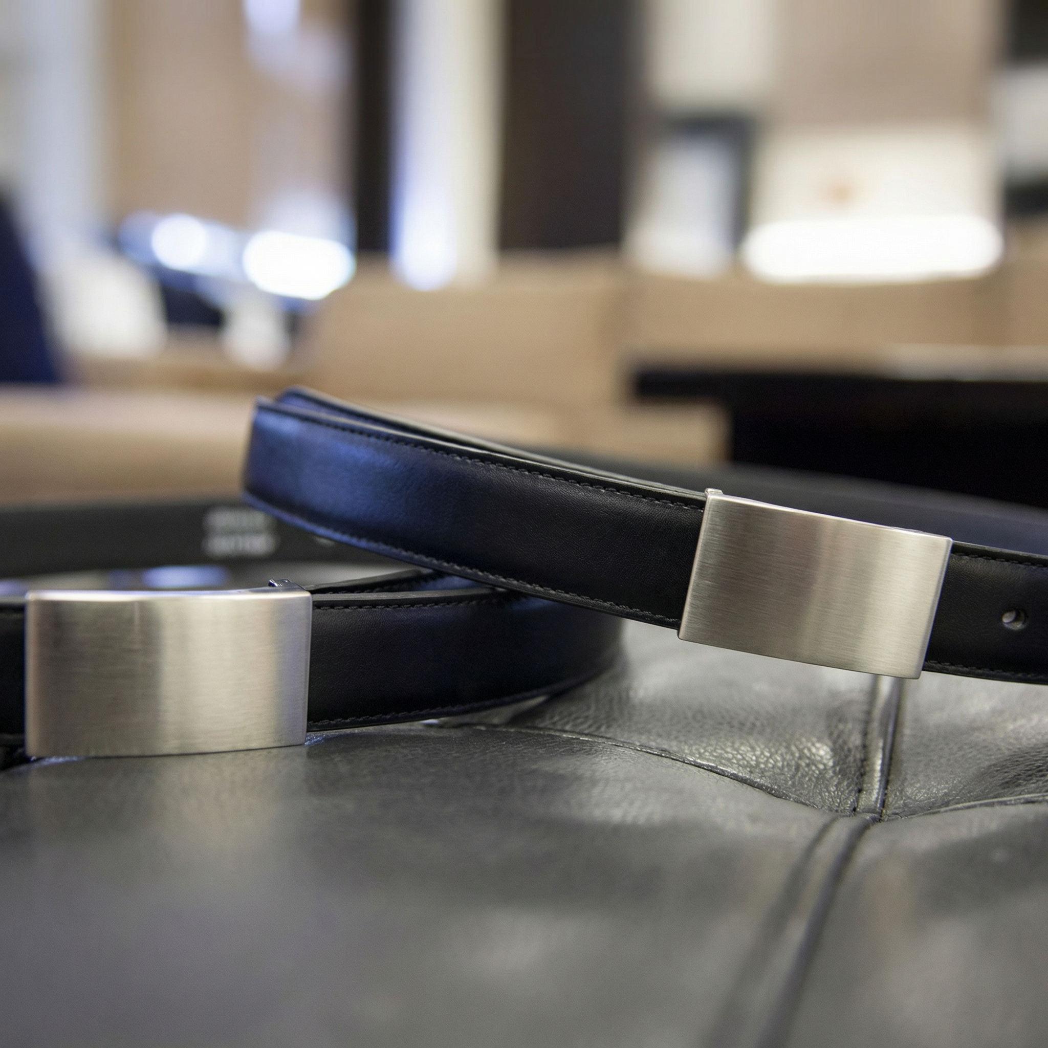 Two black leather belts featuring brushed silver buckles, angled on a dark textured surface.