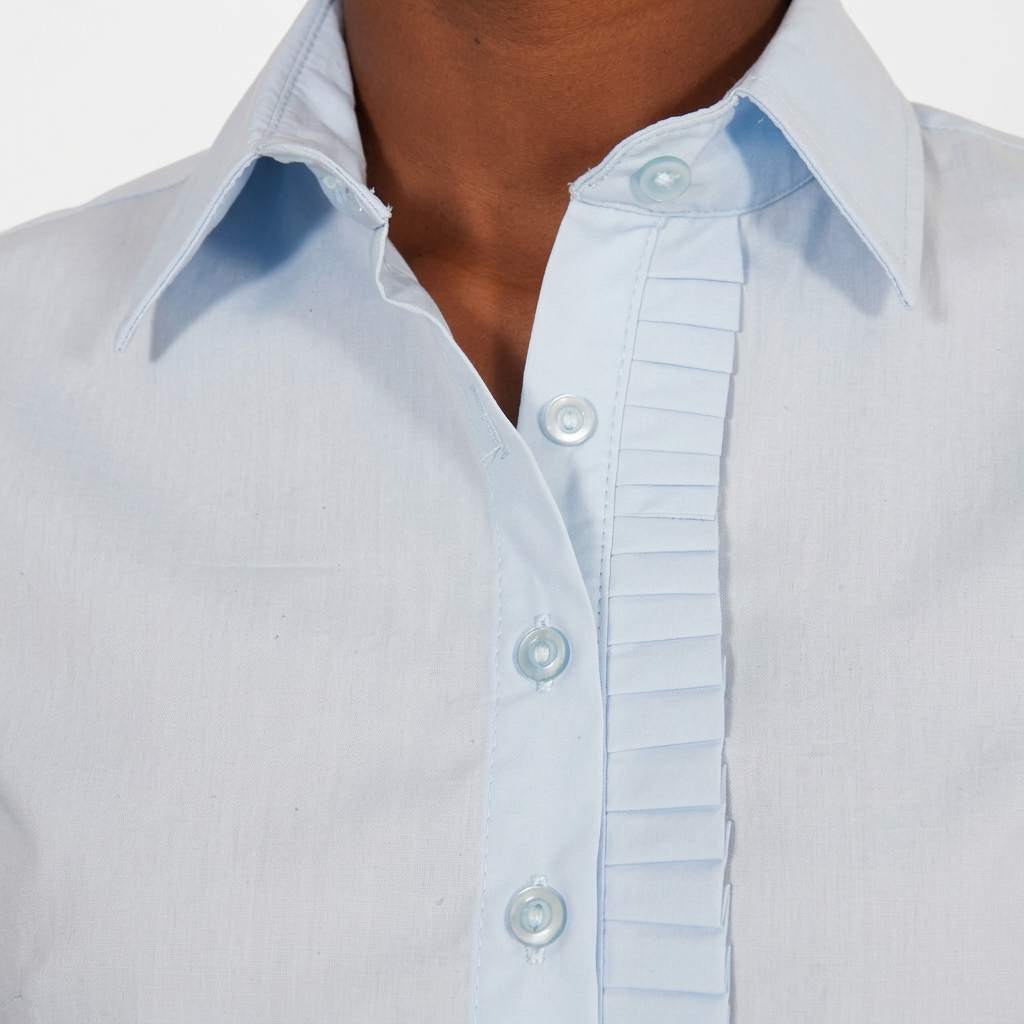 A close-up of a person wearing a light blue Domino Shirt with pleated detailing alongside the buttons. The blue shirt is neatly buttoned except for the top button, which is left open.