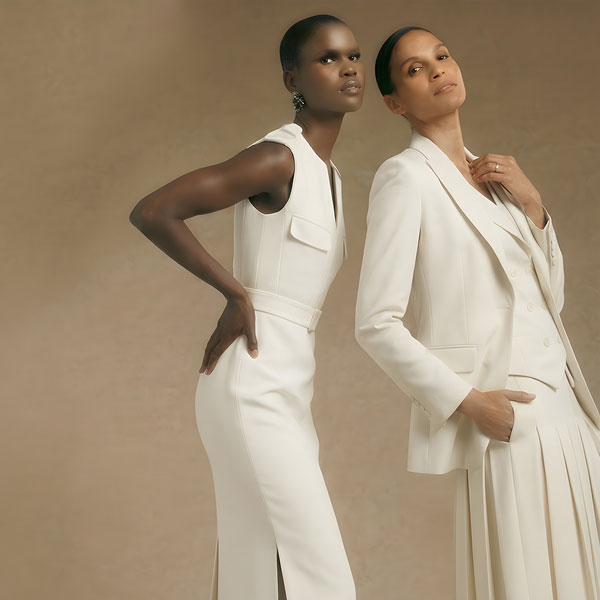 Two women in elegant cream attire. Left in sleeveless dress, hands on hips, gazing forward. Right in suit with pleated skirt, hand in pocket.